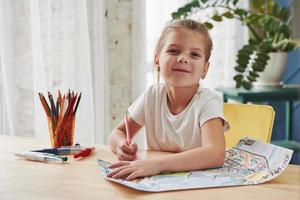met een tevreden blik in de camera kijken. schattig klein meisje op de kunstacademie tekent haar eerste schilderijen met potloden en stiften foto