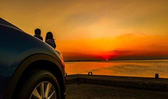 silhouet van geluk paar staan en ontspannen op het strand voor de auto met oranje en blauwe lucht bij zonsondergang. zomervakantie en reisconcept. romantisch jong stel dating aan zee. foto