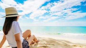 gelukkige jonge vrouw in witte overhemden en korte broeken zittend aan zandstrand. ontspannen en genieten van vakantie op tropisch paradijsstrand met blauwe lucht en wolken. meisje in zomervakantie. zomerse sferen. vrolijke dag. foto