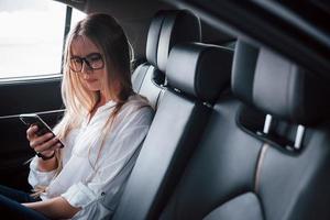 verveeld tijdens het wachten op de chauffeur. slimme zakenvrouw zit op de achterbank van de luxe auto met zwart interieur foto