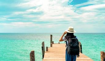 gelukkige jonge Aziatische vrouw in casual stijl mode met strohoed en rugzak. ontspan en geniet van een vakantie op het tropische paradijsstrand. meisje permanent op de houten pier van hotel in zomervakantie mode. foto