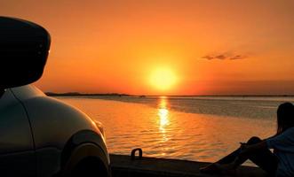 eenzame jonge vrouw draagt een pet ontspannen op het strand alleen voor de auto met oranje en blauwe lucht bij zonsondergang. zomervakantie en reisconcept. foto