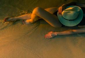 sexy Aziatische vrouw met hoed liggend op het zandstrand tijdens zonsondergang. vrouw draagt zwarte zwembroek ontspannen op het strand. zomervakantie. zomer tijd. meisje zonnebaden voor een gebruinde huid op het tropische strand van het paradijs foto