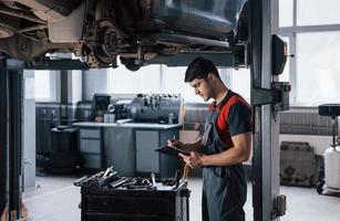 uitzicht vanaf de zijkant. man in de werkplaats in uniform met notitieblok voor zijn werk voor het repareren van kapotte auto foto