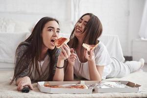 gewoon lol hebben. zusters die pizza eten terwijl ze tv kijken terwijl ze overdag op de vloer van een mooie slaapkamer liggen foto