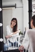 zittend op de stoel en in de spiegel kijken. jonge brunette in de kamer met witte muren en daglicht dat uit het raam komt foto