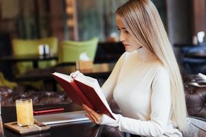 voorbereiding op examens. blond meisje las een boek met rode kaft in het restaurant met tafel en gele drank erop foto