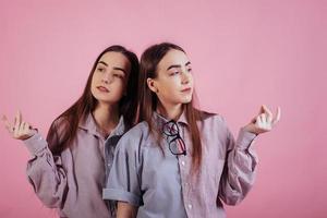 naar de zijkanten kijken. twee zussen tweeling staan en poseren in de studio met roze achtergrond foto
