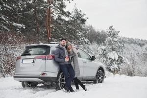 meisje zag een mooi landschap ver weg van hen. samen staan en leunend op de moderne auto in de buurt van het bos in de winter foto