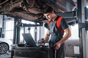 zwaar beschadigd voertuig. man in de werkplaats in uniform die laptop gebruikt voor zijn werk voor het repareren van kapotte auto foto