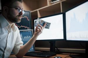 het zal zoveel kosten. bebaarde man in wit overhemd en bril houdt geld vast op kantoor met meerdere computerschermen in indexgrafieken foto