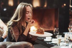 tijd voor een toetje. schattig meisje dat verse cake probeert terwijl ze in de kamer zit met een prachtige open haard foto