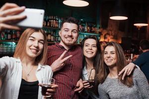 man toont gebaar van twee vingers. vrienden nemen selfie in prachtige nachtclub. met drankjes in de handen foto