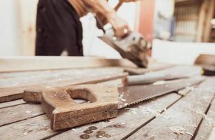 handzaag met een houten handvat op onscherpte timmerman die met elektrische houtschaafmachine werkt. timmerman gereedschap. handzaag en zaagsel op houten tafel bij outdoor workshop. ambachtsman die houtwerk maakt. handwerkconcept. foto