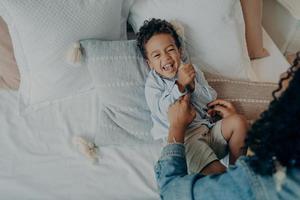 lieve opgewonden jongen van gemengd ras die op bed ligt en lacht tijdens het spelen met zijn liefhebbende moeder foto