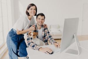 afbeelding van een gelukkige brunette glimlachende vrouw leunt op de schouders van de echtgenoot die toetsenborden op de computer, op internet surft en een nieuw appartement zoekt om te kopen op de website, poseert in een ruime kamer met een huishond foto