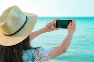 jonge aziatische vrouw draagt hoed gebruik smartphone die foto neemt op tropisch strand. zomervakantie op tropisch paradijsstrand. gelukkig hipster meisje reizen op vakantie. vrouw genieten en ontspannen leven. zomerse sferen.