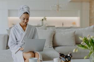 horizontaal schot van mooie jonge vrouw gekleed in badjas en handdoek na het douchen zit op een comfortabele bank in de woonkamer werkt op laptopcomputer heeft eigen beautyblogtypes feedback aan volgers foto