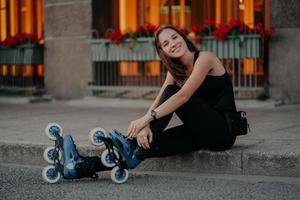 aangenaam ogende jonge europese vrouw draagt rolschaatsen neemt pauze na het rijden poses buiten gekleed in zwarte activewear heeft een gelukkige glimlach op het gezicht. mensen recreatie hobby en lifestyle concept foto
