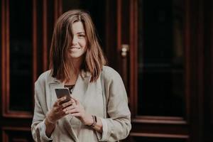 aantrekkelijke jonge glimlachende vrouwelijke blogger sms't berichten naar volgers, heeft vrije tijd, is in een goed humeur, draagt een witte modieuze regenjas, staat buiten bij deuren. communicatie concept foto
