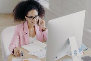 foto van gelukkige vrouw met krullend haar draagt optische bril, roze jas, kijkt aandachtig naar computerscherm, zit aan desktop met geopend notitieblok, belt iemand via smartphone, heeft een positieve glimlach