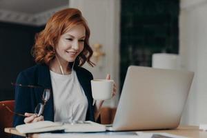 jonge vrouwelijke lerares geeft online les, praat met leerlingen via laptop, organiseert videoconferentie, geeft les aan studenten, drinkt aromatische koffie, houdt een bril in de hand, is formeel gekleed foto