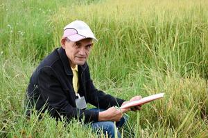 portret van aziatische senior vrijwilliger die wit papier en pen in de hand houdt, zit op gras in de buurt van rijstveld, concept voor een gelukkig leven na pensionering van behulpzame en vriendelijke aziatische mensen. foto