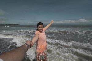 mooie gelukkige jonge Spaanse vrouw die de hand van haar partner vasthoudt op het strand, gekleed in een roze jurk tijdens een zonnige ochtend foto