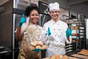 portret van twee professionele koks in uniform die naar de camera kijken met een vrolijke, trotse glimlach en duim omhoog met brood in de keuken. een partner van commerciële banketbakkerij en verse bakkerijkeuken. foto