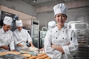 jonge mooie aziatische vrouwelijke chef-kok in wit kookuniform kijkt naar camera, gekruiste armen en vrolijke glimlach met voedsel professionele bezetting, commerciële gebak culinaire banen in de keuken van een restaurant. foto