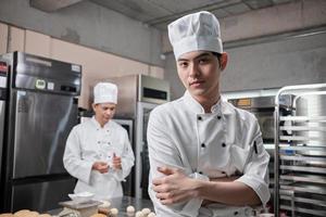 portret van een jonge Aziatische mannelijke chef-kok in wit kookuniform kijkend naar de camera, armen gekruist met vertrouwen, voedingsmiddelen professionele bezetting, commerciële gebak culinaire banen in de keuken van een restaurant. foto