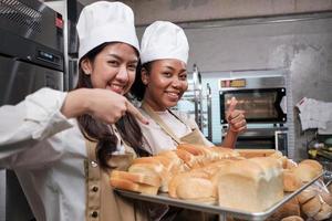 portret van twee jonge vrouwelijke chef-koks in kookuniform die naar de camera kijken met een vrolijke glimlach en trots met een dienblad met brood in de keuken. vriend en partner van bakkerijproducten, dagelijks verse bakkerijberoep. foto