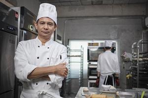 professionele senior Aziatische mannelijke chef-kok in wit kookuniform, voedselbezetting kijkt naar camera, armen gekruist, expertise in commerciële culinaire banketbakkerijen, cateringrestaurantkeuken. foto