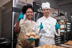 portret van twee professionele koks in uniform die naar de camera kijken met een vrolijke, trotse glimlach en duim omhoog met brood in de keuken. een partner van commerciële banketbakkerij en verse bakkerijkeuken. foto