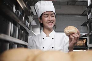 jonge Aziatische vrouwelijke chef-kok in wit koksuniform en hoed met dienblad met vers lekker brood met een glimlach, kijkend naar haar broodje, blij met zijn gebakken voedselproducten, professionele baan bij roestvrijstalen keuken. foto