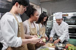 hobbykeukencursus, senior mannelijke chef-kok in kookuniform leert jonge kooklesstudenten appels te schillen en hakken, ingrediënten voor banketbakkerswaren, fruittaarten in de roestvrijstalen keuken van het restaurant. foto