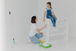 blije brunette moeder kijkt naar kind, verf meubels in modern appartement, poseert in ruime lege kamer met witte muren, bezig met huisrenovatie. familie-, reparatie- en schilderconcept foto