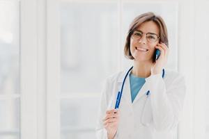 positieve vrouwelijke arts raadpleegt patiënt via mobiele telefoon, kijkt graag naar de camera, houdt een pen vast, draagt een witte jas, poseert in het medische kantoor, poseert bij het raam. gezondheidszorg en overleg concept. foto