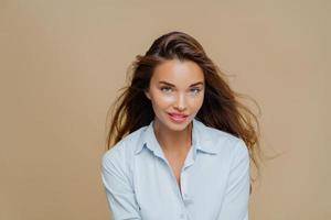 mooie duizendjarige vrouw kijkt met een tevreden uitdrukking naar de camera, heeft lang haar, gekleed in een stijlvol shirt, heeft natuurlijke schoonheid, modellen tegen een bruine achtergrond, draagt make-up, praat levendig foto