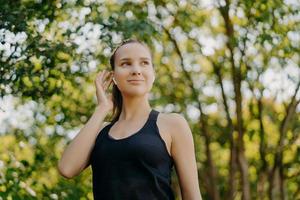 horizontaal schot van tevreden jonge vrouw houdt hand op oordopje luistert favoriete muziek kijkt weg heeft tevreden gezichtsuitdrukking gekleed in actieve kleding geniet zomerochtend buiten heeft cardiotraining foto