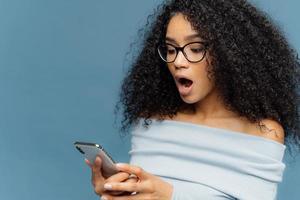 geschokte afro-amerikaanse vrouw krijgt bericht van ex, hapt naar adem van verbazing en opwinding, leest verbluffend nieuws op mobiele telefoon, heeft krullend borstelig haar, draagt trui, geïsoleerd over blauwe studiomuur foto