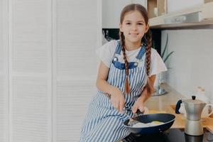 binnenopname van een prettig uitziend zevenjarig meisje dat bezig is met het bakken van eieren, houdt de pan vast en zit bij het fornuis, gekleed in een schort, kijkt positief naar de camera, schept op dat ze kan koken, poseert in de moderne keuken foto