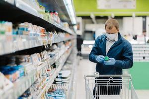 horizontaal schot van volwassen man draagt beschermend masker, leest het etiket van het product, doet boodschappen tijdens de uitbraak van het coronavirus, koopt het noodzakelijke voedsel in de plaatselijke winkel. mensen, virus, ziekte, aankoopconcept foto