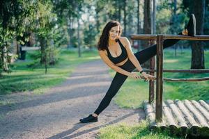 flexibele jonge krullende vrouw draagt zwarte top legging en sneakers, strekt de benen, sport tijdens de zomerdag in de open lucht, poseert tegen groene bomen. mensen, flexibiliteit en bewegingsconcept foto