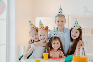 groep schattige kinderen dragen feestmutsen, omhelzen en hebben plezier, vieren verjaardag, poseren in een versierde kamer, verzamelen zich in de buurt van een feesttafel, veel plezier knuffelen en kijken graag naar de camera foto