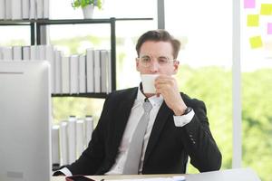 knappe zakenman in zwart pak draagt een bril die blij is met relaxtime en koffie drinkt aan de computerbalie op kantoor foto