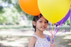 schattig klein meisje met kleurrijke ballonnen in de wei tegen de blauwe lucht en de wolken, handen spreidend. foto