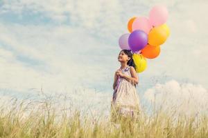 schattig klein meisje met kleurrijke ballonnen in de wei tegen de blauwe lucht en de wolken, handen spreidend. foto