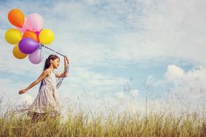 schattig klein meisje met kleurrijke ballonnen in de wei tegen de blauwe lucht en de wolken, handen spreidend. foto