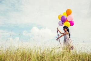 schattig klein meisje met kleurrijke ballonnen in de wei tegen de blauwe lucht en de wolken, handen spreidend. foto